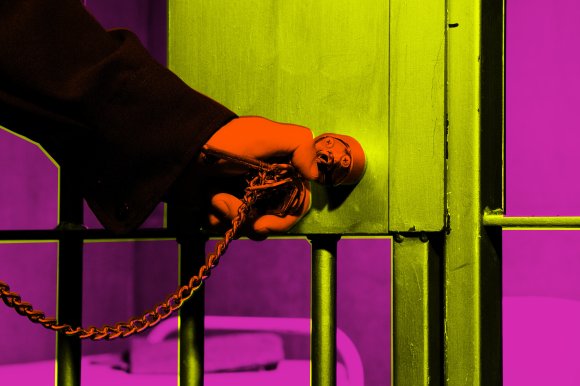 A hand holding a key inserted into a lock on a metal prison door with vertical bars. The hand and key are highlighted in orange, the door is yellow-green, and the background is purple, creating a vivid, high-contrast color effect.