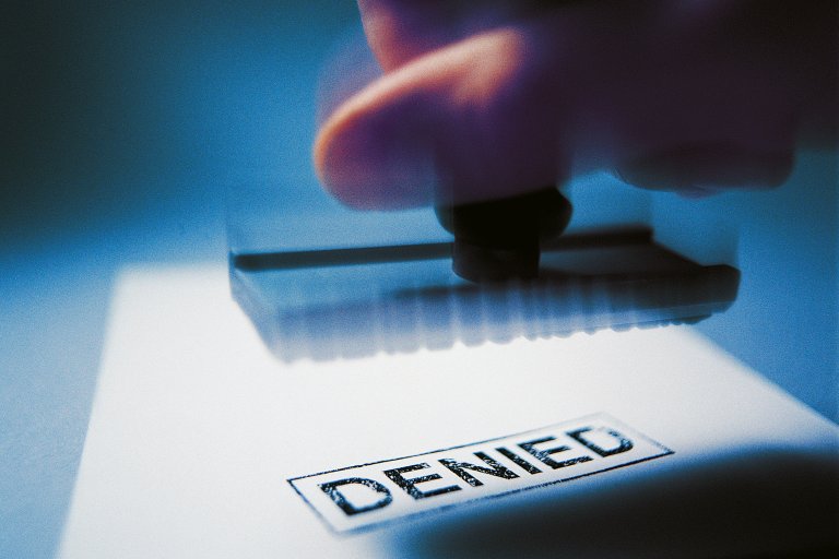 A close-up of a hand holding a rubber stamp that has just stamped the word "DENIED" in black ink on a white surface. The image has a blue tint and a slightly blurred effect.