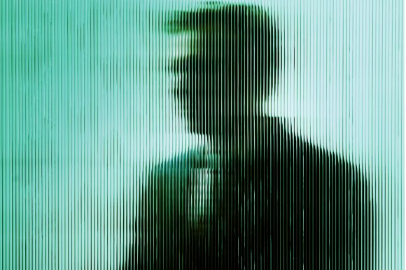 Silhouette of a person viewed through vertically ribbed translucent glass, creating a distorted and blurred effect. The figure appears to be facing left, with short hair and wearing a dark top. The background is light blue-green.