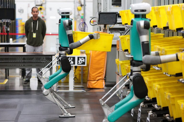 Two teal and silver robots with white heads are handling yellow bins in an industrial setting, likely a warehouse or sorting facility. One robot is holding a yellow bin, while the other is positioned near a row of yellow bins on shelves. A person in a green hoodie and gray pants stands in the background observing the robots. The environment includes industrial equipment, tables, and various cables.