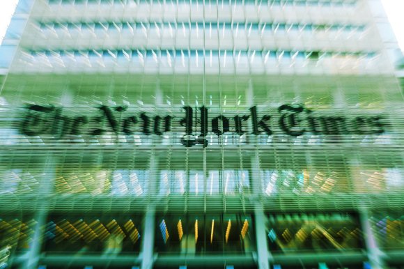 The image shows the exterior of the New York Times building with its name prominently displayed on the glass facade. The photo has a motion blur effect, creating a dynamic and slightly distorted appearance of the building and its signage.