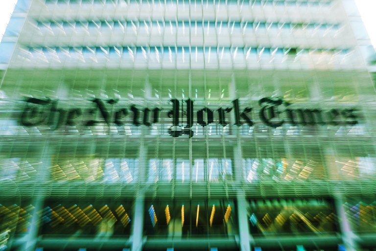 The image shows the exterior of the New York Times building with its name prominently displayed on the glass facade. The photo has a motion blur effect, creating a dynamic and slightly distorted appearance of the building and its signage.