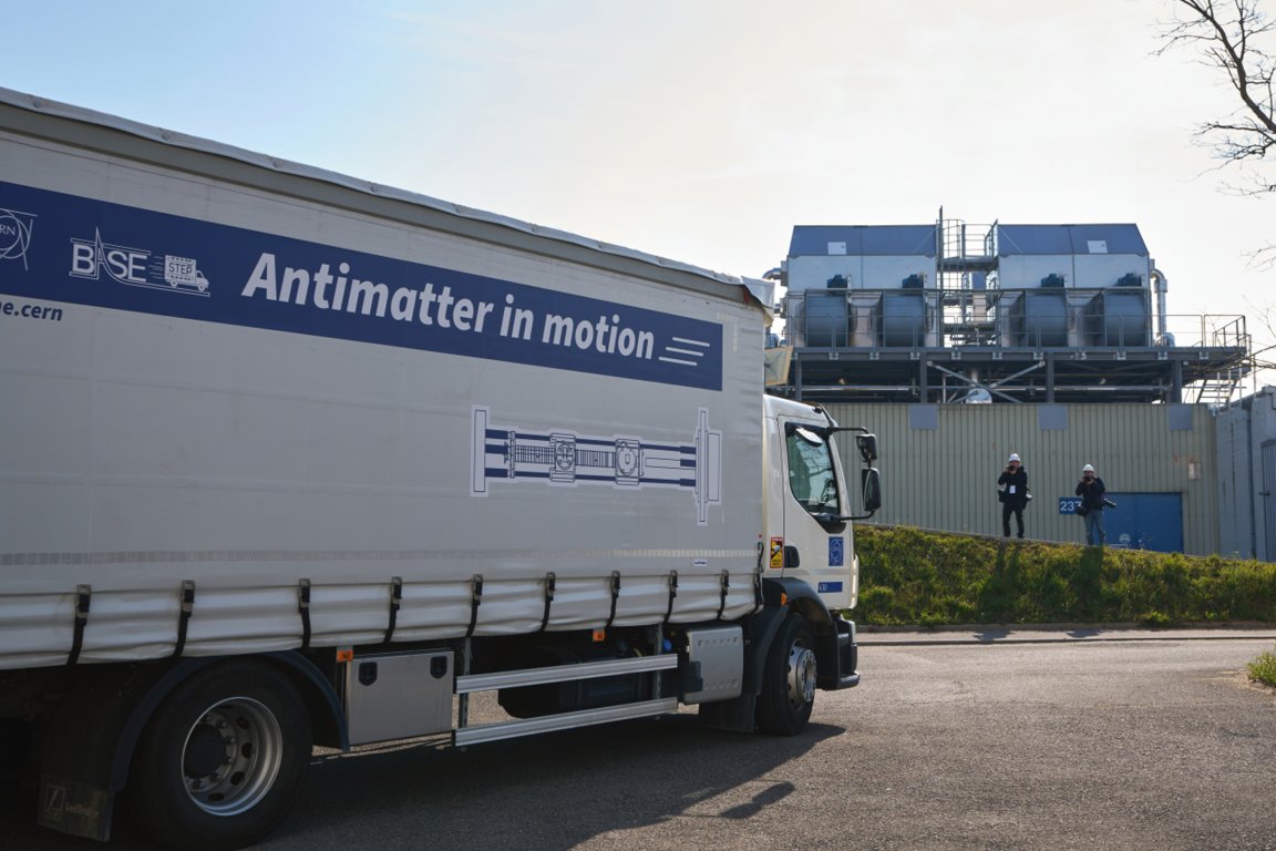 A large white truck with the phrase "Antimatter in motion" printed on its side is parked on a paved area. In the background, there is an industrial building with large ventilation units on the roof. Two people wearing helmets and dark clothing are standing near the building, one holding a camera. The scene is outdoors on a clear day.