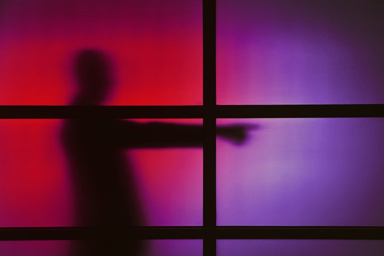 Silhouette of a person behind a frosted glass panel divided by black horizontal and vertical bars, with the person pointing to the right. The background features a gradient of red and purple hues.