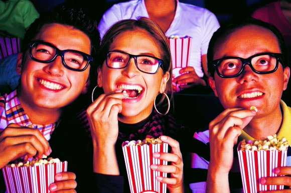 Three people wearing 3D glasses are sitting close together, smiling and eating popcorn from red and white striped containers. The person in the middle is a woman with hoop earrings, while the two on either side are men. The background is dark, suggesting they are in a movie theater.