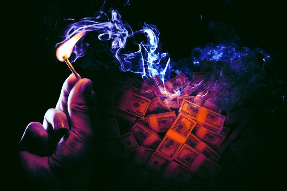 A hand holding a lit matchstick near a pile of burning US dollar bills, with blue and purple smoke rising against a dark background.