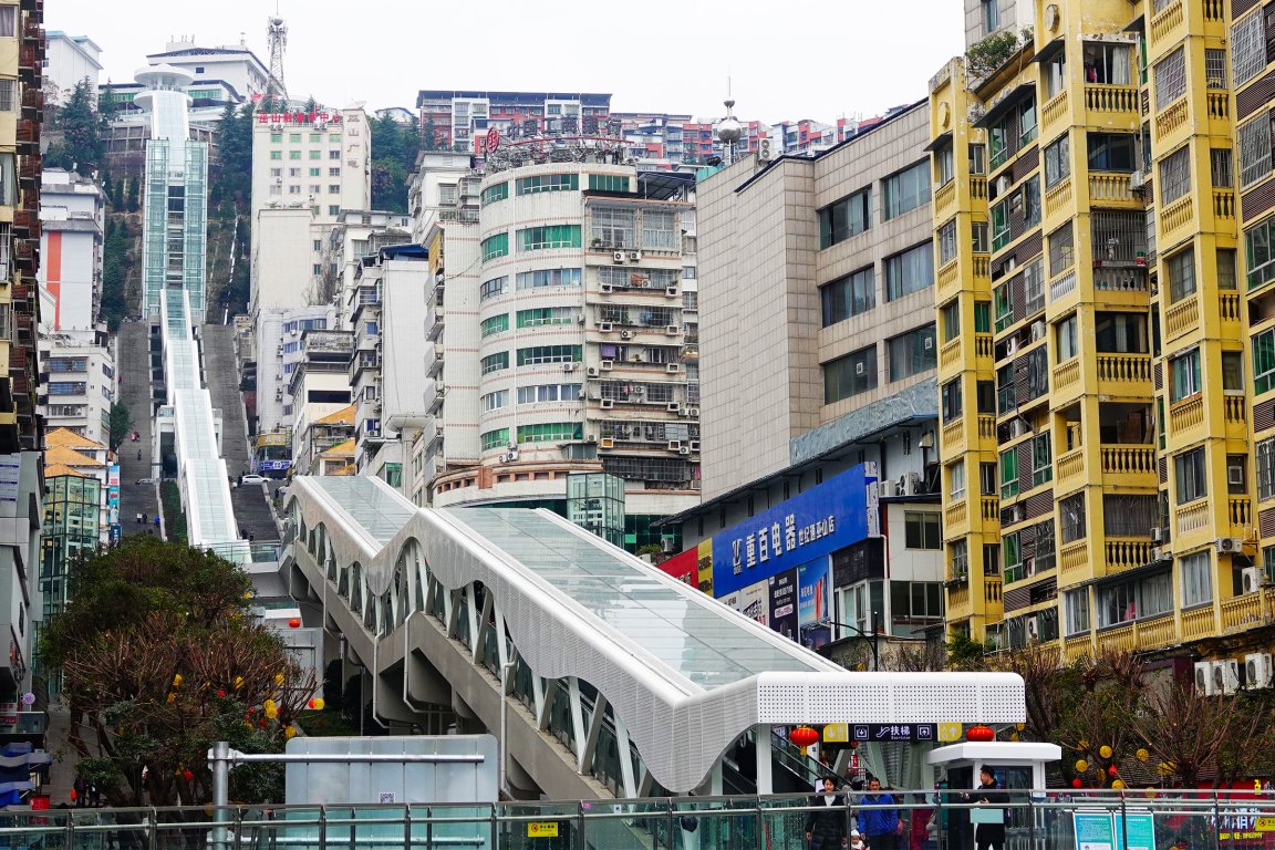 A long outdoor escalator with a glass roof runs uphill between densely packed urban buildings. The escalator is covered with a white, wavy metal and glass canopy. Surrounding the escalator are tall residential and commercial buildings, including a yellow apartment building on the right and a rounded, multi-story building with green windows in the center. The area appears busy with a few people visible near the escalator entrance. The scene is set in a city with a hilly terrain.