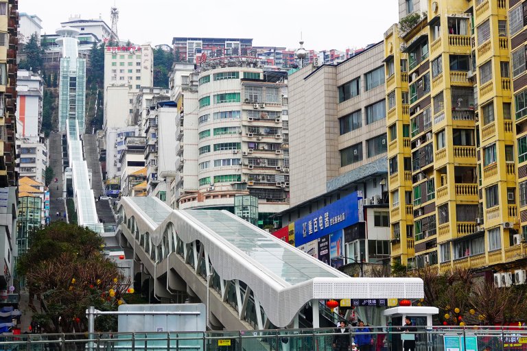A long outdoor escalator with a glass roof runs uphill between densely packed urban buildings. The escalator is covered with a white, wavy metal and glass canopy. Surrounding the escalator are tall residential and commercial buildings, including a yellow apartment building on the right and a rounded, multi-story building with green windows in the center. The area appears busy with a few people visible near the escalator entrance. The scene is set in a city with a hilly terrain.
