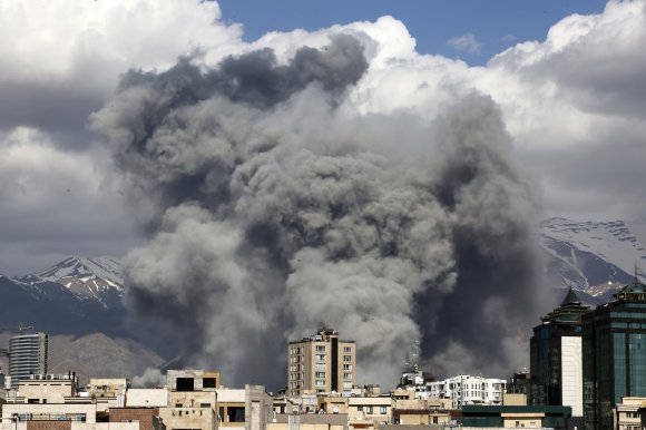 Thick, dark smoke billows over a cityscape with mid-rise buildings, set against a backdrop of snow-capped mountains and a partly cloudy sky.