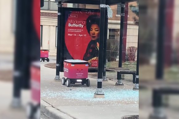 A small red delivery robot with the label "COCO" is at a bus stop with shattered glass scattered on the ground around it. Behind the robot, there is a poster advertising Puccini's opera "Madama Butterfly," showing a woman in traditional attire with the dates Mar 14 - Apr 12. The bus stop has black metal poles and a bench.