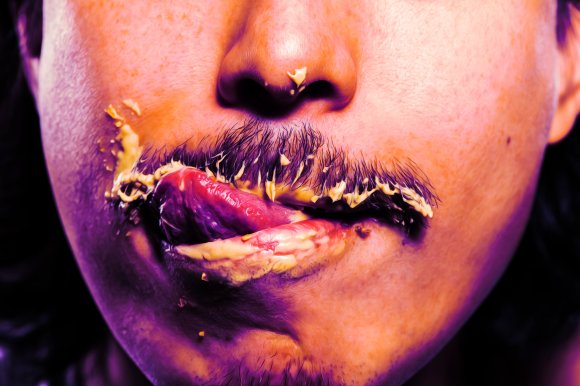 Close-up of a person's lower face with a mustache and some facial hair, smeared with a creamy yellow substance around the mouth and on the upper lip, with the tongue partially sticking out and touching the lips. The skin has a warm tone with a slight purple hue in the shadows.