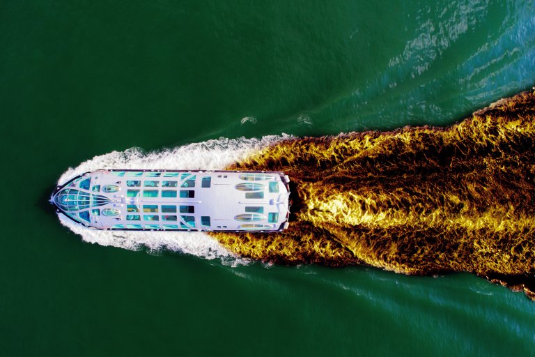 A white boat with a glass-covered upper deck is moving through green water, creating a striking wake with a mix of white foam and dark brown water trailing behind it. The image is taken from directly above, showing the boat's streamlined shape and the vivid contrast between the wake and the surrounding water.