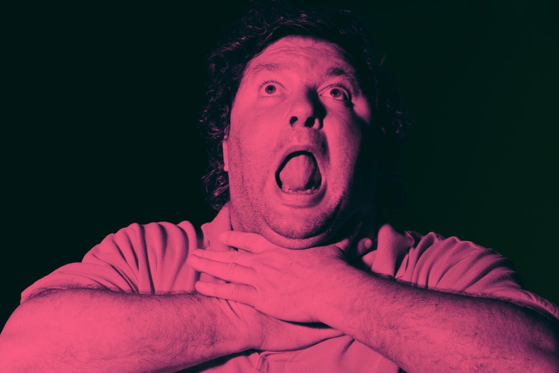 A man with curly hair is shown with his mouth open wide and eyes looking upward, clutching his throat with both hands as if choking or struggling to breathe. The image has a strong pinkish-red tint and a dark background.