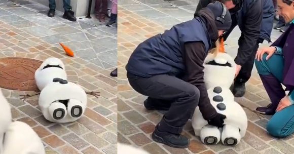 A robot version of Olaf from "Frozen" is lying on its back on a cobblestone street. Nearby, a person wearing a dark jacket, gloves, and a beanie is crouching and holding the snowman, while another person kneels beside them. Several people stand around observing.