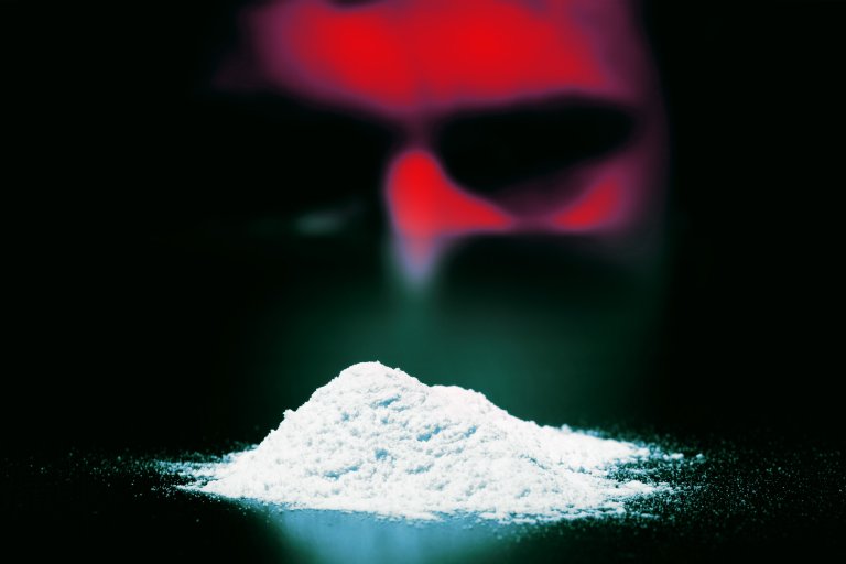 A small mound of fine white powder on a dark surface with a blurred red and black face in the background.