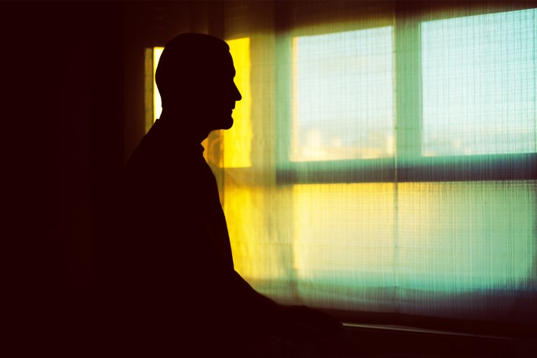 Silhouette of a person sitting indoors near a window with sheer curtains, illuminated by soft yellow and green light filtering through the window.