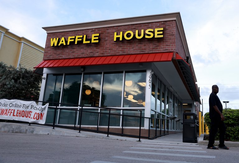 Waffle House restaurant with a brick facade and large yellow letters spelling "WAFFLE HOUSE" above a red awning. The building has large glass windows reflecting the sky and interior lights. A man dressed in black is walking near the entrance, and a banner on the left side advertises online ordering for pickup.