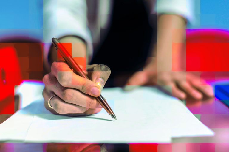 A close-up of a hand holding a brown pen poised to write on a blank sheet of white paper. The person is wearing a ring on their ring finger and a light-colored long sleeve shirt. The background is blurred with hints of blue and red colors.