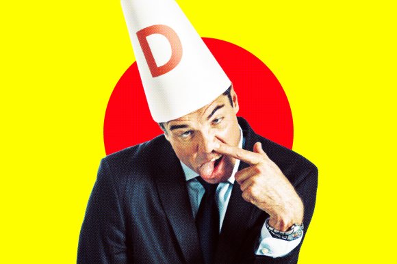 A man in a black suit and tie wearing a white dunce cap with a red letter "D" on it. He is making a silly face, sticking out his tongue and picking his nose with his right hand. The background is bright yellow with a large red circle behind his head.