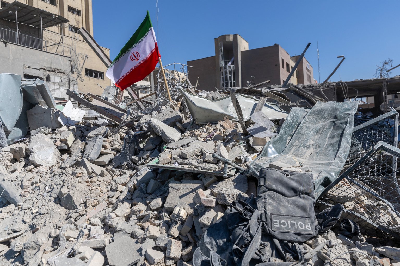 Rubble and debris from a destroyed building with a dusty police vest lying on top. An Iranian flag is planted in the rubble, and damaged buildings are visible in the background under a clear blue sky.