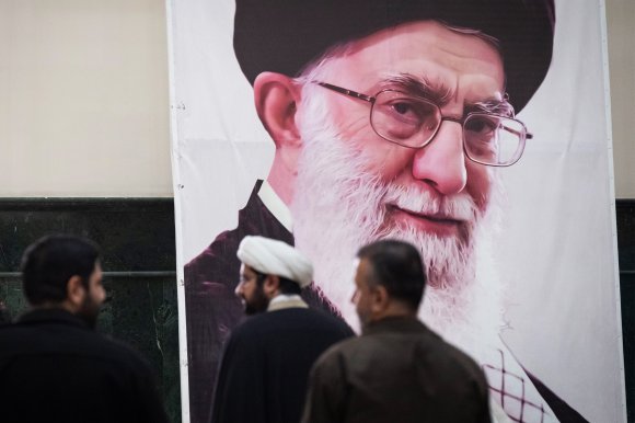 A large portrait of former Iranian leader Ali Khamenei, with a white beard, glasses, and a black turban, displayed on a wall. In front of the portrait, three men are standing and looking towards it, one of whom is wearing a white turban and traditional clothing. The setting appears to be indoors.