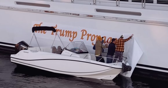 Three people on a small white motorboat are applying a large white banner with orange text to the side of a larger white boat.