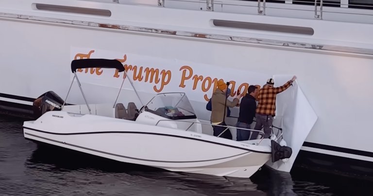 Three people on a small white motorboat are applying a large white banner with orange text to the side of a larger white boat.