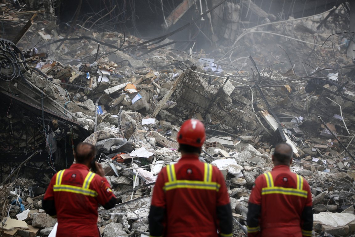 Three rescue workers in red uniforms with yellow reflective stripes stand facing a large pile of rubble and debris from a collapsed structure. The scene is chaotic with broken concrete, twisted metal rods, and scattered materials. One worker wears a red helmet, while another is using a phone. Dust and smoke create a hazy atmosphere.