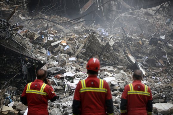 Three rescue workers in red uniforms with yellow reflective stripes stand facing a large pile of rubble and debris from a collapsed structure. The scene is chaotic with broken concrete, twisted metal rods, and scattered materials. One worker wears a red helmet, while another is using a phone. Dust and smoke create a hazy atmosphere.