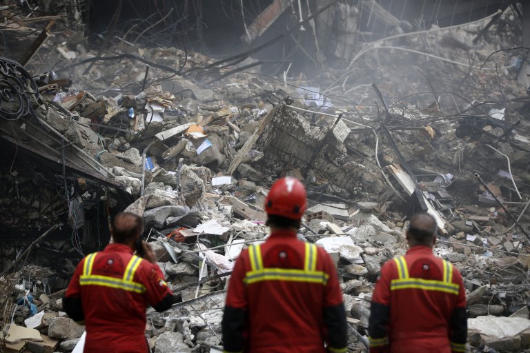Three rescue workers in red uniforms with yellow reflective stripes stand facing a large pile of rubble and debris from a collapsed structure. The scene is chaotic with broken concrete, twisted metal rods, and scattered materials. One worker wears a red helmet, while another is using a phone. Dust and smoke create a hazy atmosphere.