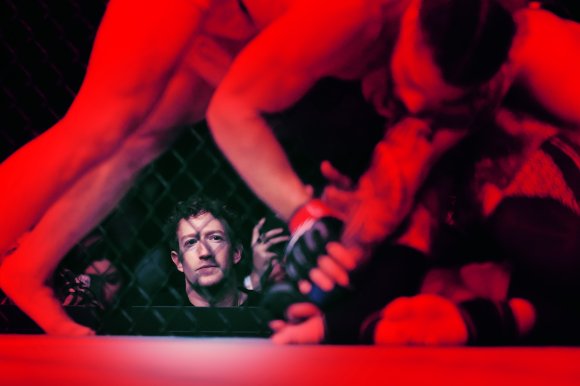 Mark Zuckerberg watches intently through a chain-link fence at a mixed martial arts fight inside a cage, where two fighters are engaged on the ground under red lighting.