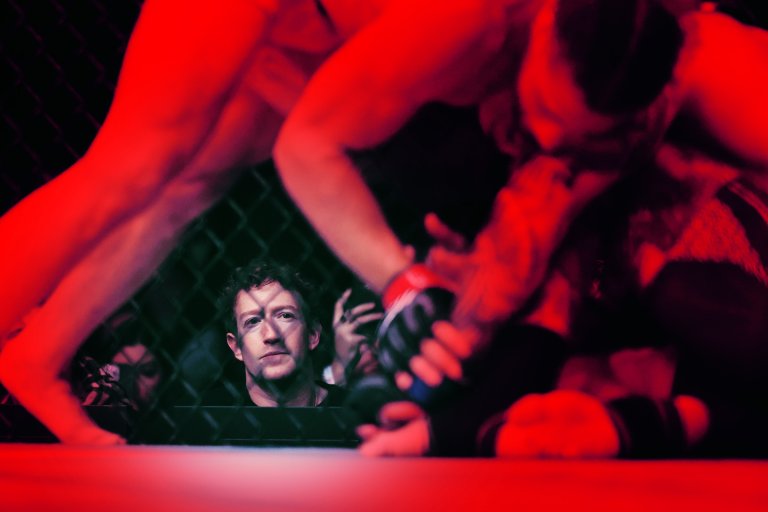 Mark Zuckerberg watches intently through a chain-link fence at a mixed martial arts fight inside a cage, where two fighters are engaged on the ground under red lighting.