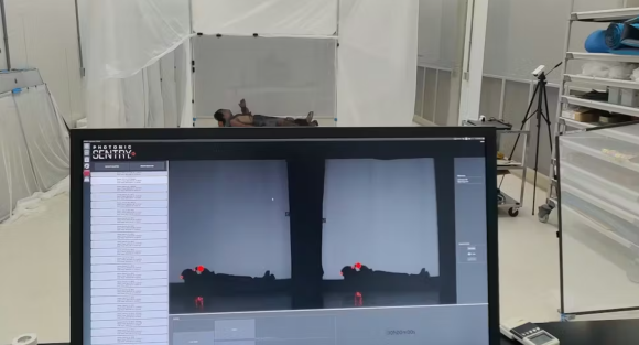 A person is lying on their back on a flat surface in a white room with translucent curtains. In the foreground, there is a computer monitor displaying a software interface labeled "Photonic Sentry," showing two dark silhouettes of the person with red markers on their head and feet, likely indicating tracking points. The room contains various equipment, including a tripod with a camera or sensor and storage shelves with rolled mats and other items.