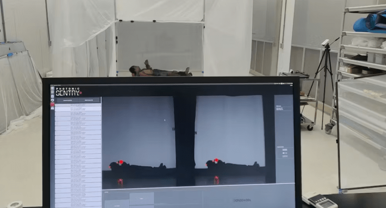 A person is lying on their back on a flat surface in a white room with translucent curtains. In the foreground, there is a computer monitor displaying a software interface labeled "Photonic Sentry," showing two dark silhouettes of the person with red markers on their head and feet, likely indicating tracking points. The room contains various equipment, including a tripod with a camera or sensor and storage shelves with rolled mats and other items.