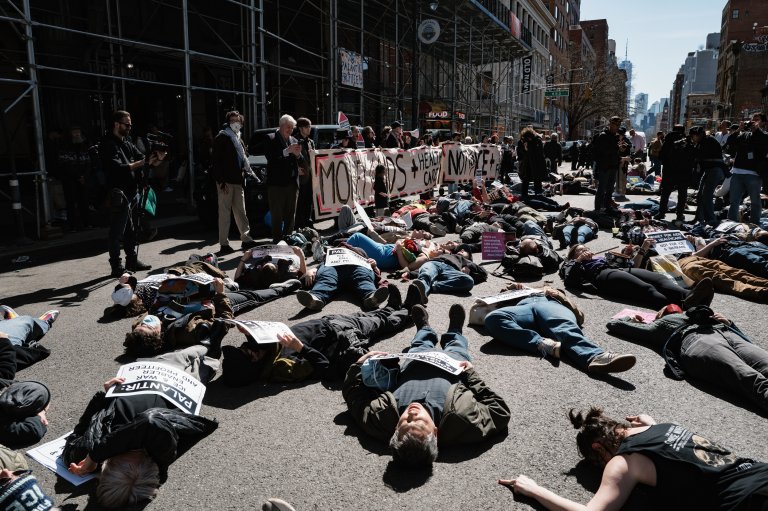 A large group of people participating in a protest, many lying down on the street holding signs with messages such as "PALANTIR: ICE, WAR PROFITEER AND PROTECTOR" and "MONEY FOR AIDS HEALTHCARE NOT FOR WAR & PROFIT." Others stand behind a large banner that reads "MOTHERS + HEALTH CARE NOT WAR." The scene takes place on a city street with scaffolding on one side and tall buildings in the background. Some individuals are taking photos or filming the event.