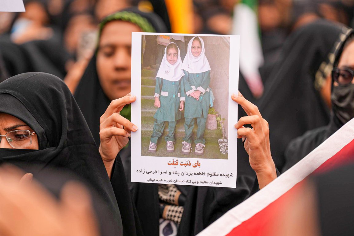 A person wearing a black hijab and glasses is holding a photo of two young girls dressed in matching green school uniforms and white headscarves. The photo includes text in Persian below the image of the girls. The background shows a crowd of people, many also wearing black clothing and head coverings.