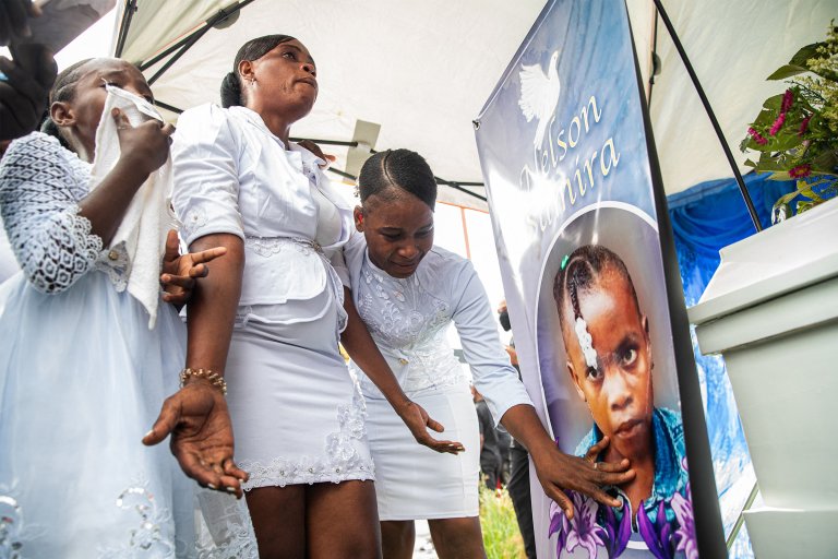 Three women dressed in white are mourning beside a large banner with a photo of a young girl and the name "Nelson Samira." One woman is wiping tears with a cloth, another looks upward with a somber expression, and the third is touching the banner near the girl's face.