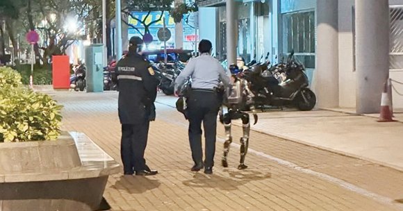Two security personnel are walking on a paved sidewalk at night, escorting a humanoid robot. The robot has a metallic frame with visible joints and a backpack-like component. The scene is illuminated by streetlights, with motorcycles parked along the side and some greenery on the left.