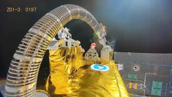 A close-up view of a spacecraft component covered in gold foil with a flexible, ribbed metallic tube arching over it. The background is the blackness of space, and part of a solar panel with circuitry is visible on the right side. The image has a timestamp or code "Z01-3: 0197" in the upper left corner.