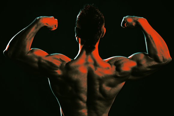 Muscular man flexing his biceps with his back facing the camera, highlighted by dramatic red and teal lighting against a dark background.