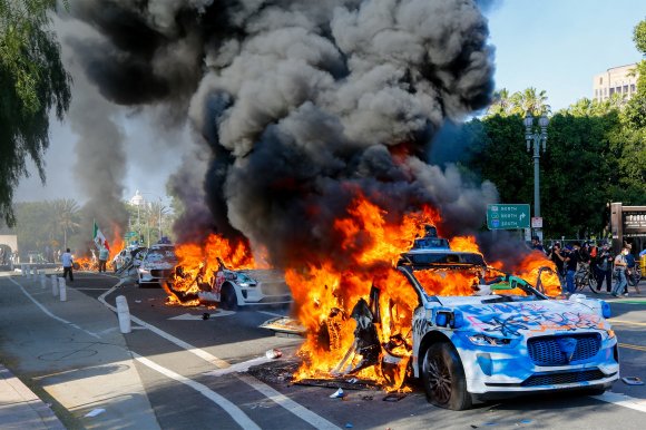 Several Waymo vehicles are shown covered in graffiti are engulfed in large flames and thick black smoke on a city street, with people and photographers observing from the sidewalk. The scene appears chaotic and intense, with bright orange fire contrasting against the blue sky and green trees in the background.