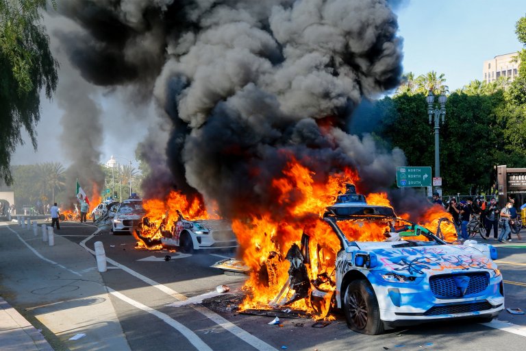 Several Waymo vehicles are shown covered in graffiti are engulfed in large flames and thick black smoke on a city street, with people and photographers observing from the sidewalk. The scene appears chaotic and intense, with bright orange fire contrasting against the blue sky and green trees in the background.
