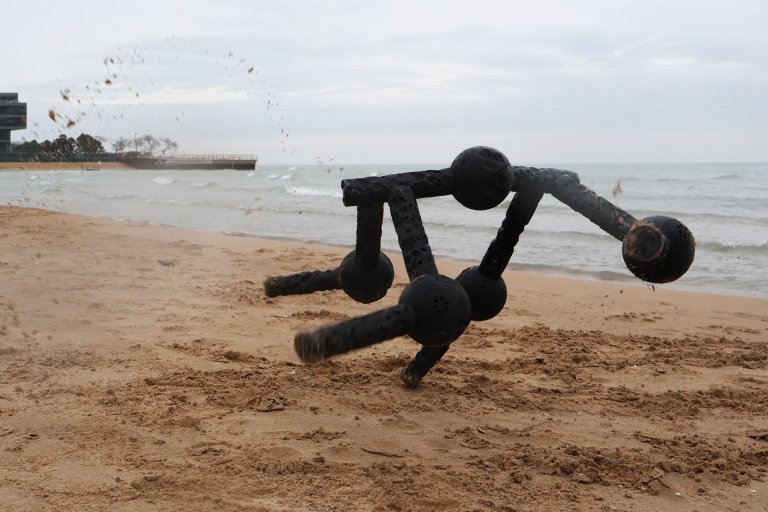 A black robot made of interconnected segments is positioned on a sandy beach near the water. The robot is in motion, kicking up sand in an arc behind it. The background shows a calm sea under a cloudy sky, with a distant pier and some trees on the left side.