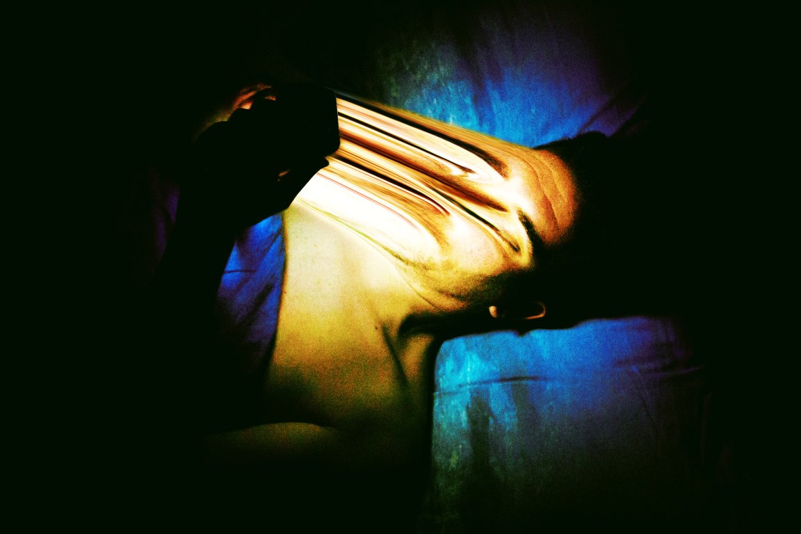 A person lying down on a blue surface with their face distorted in a stretched, abstract manner. The lighting highlights the face and upper body, creating a dramatic contrast with the dark background. The distortion gives the image a surreal and unsettling effect.