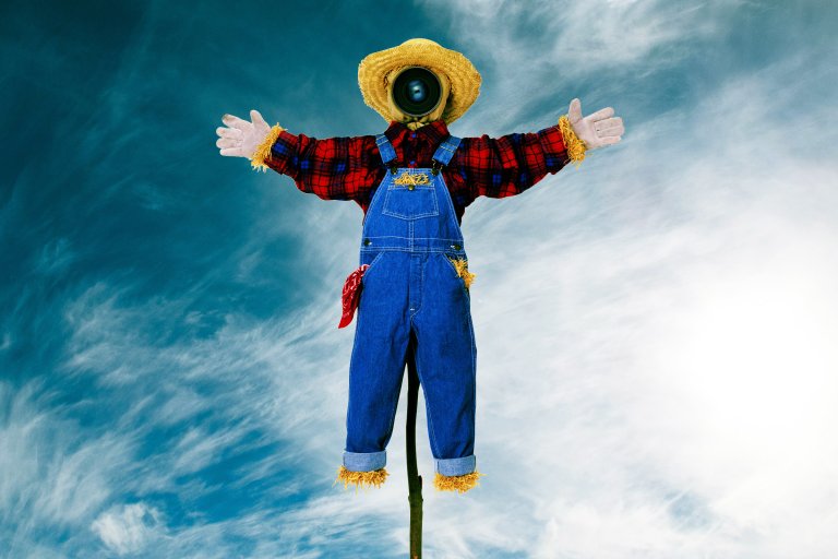 A scarecrow dressed in blue denim overalls and a red plaid shirt with straw accents at the cuffs and ankles. It wears a straw hat and has a camera lens for a face. A red bandana is tucked into the overalls' pocket. The scarecrow is set against a partly cloudy blue sky.