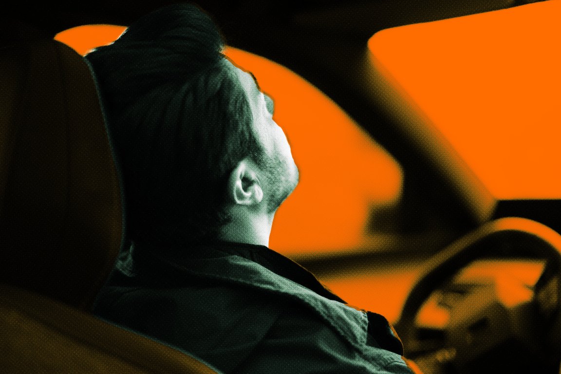 Side view of a man with dark hair sitting in the driver's seat of a car, looking upwards. The background outside the car windows is bright orange, creating a high-contrast effect with the dark interior and the man's silhouette.