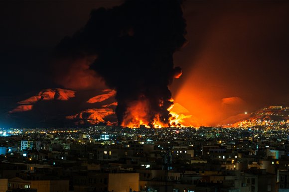 A large fire with intense orange flames and thick black smoke rising into the night sky, illuminating nearby mountains and a densely built urban area in the foreground. The fire's glow casts a dramatic light over the surrounding landscape.
