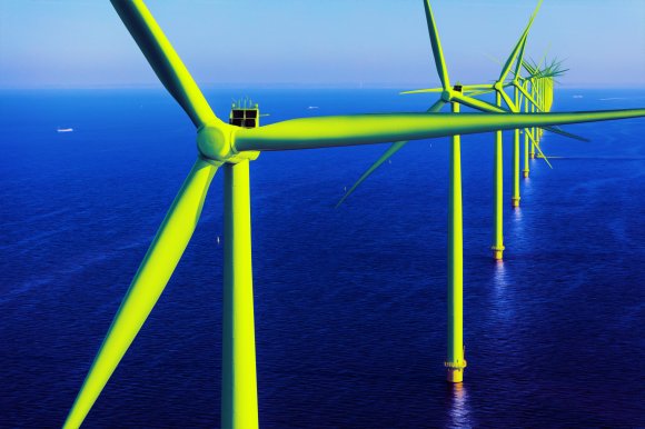 A long row of offshore wind turbines extends into the distance over a deep blue sea under a clear sky. The turbines are painted bright yellow-green, with large blades and tall cylindrical towers anchored in the water. Several ships are visible far in the background on the horizon.