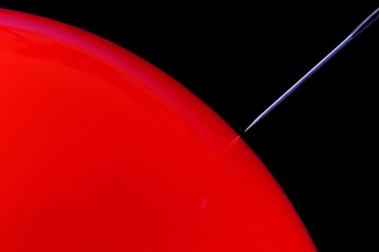 A close-up of a sharp needle piercing the surface of a red balloon against a black background. The needle is thin and metallic, positioned at an angle, just touching the balloon's surface. The balloon's red surface is smooth and glossy.