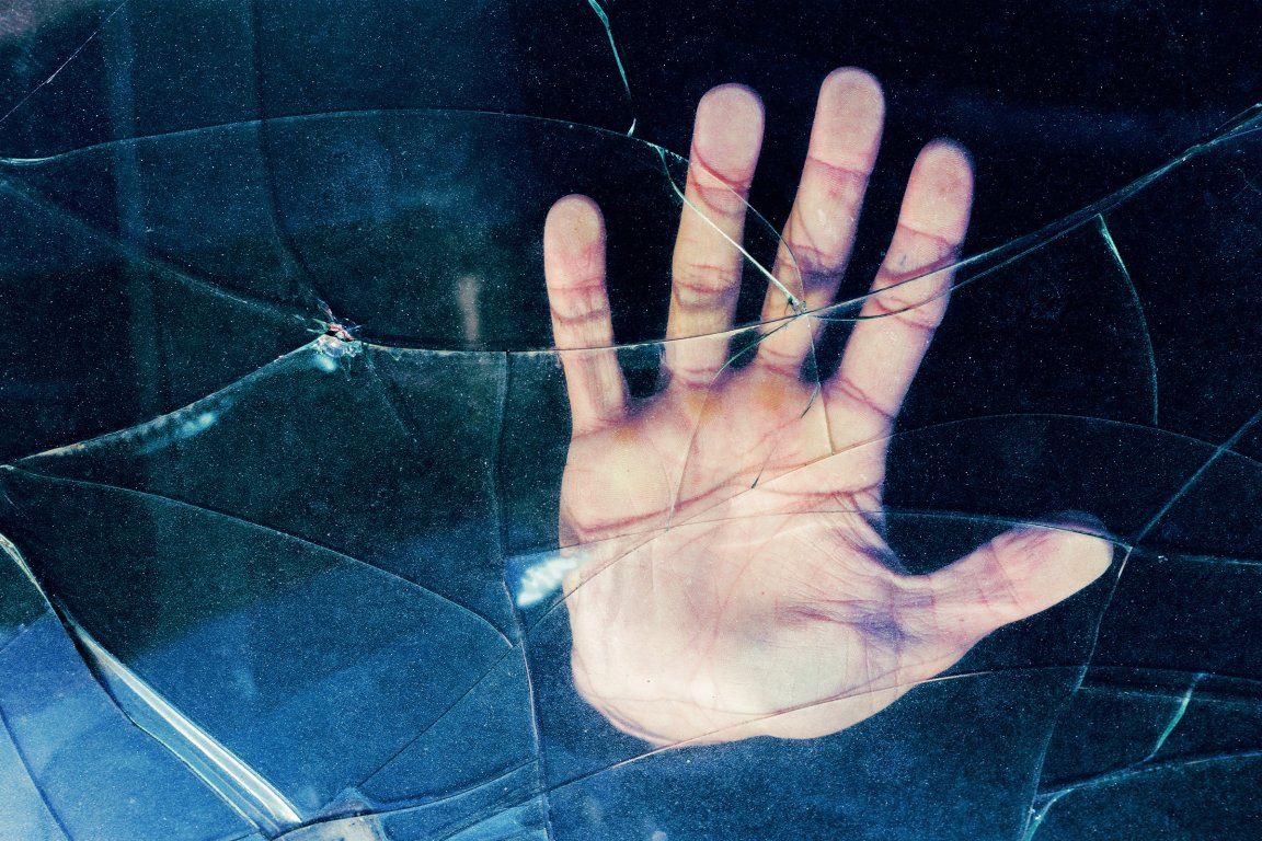 A close-up of a human hand pressed against a surface of cracked glass, with the cracks radiating around the hand. The background is dark, highlighting the hand and the broken glass.